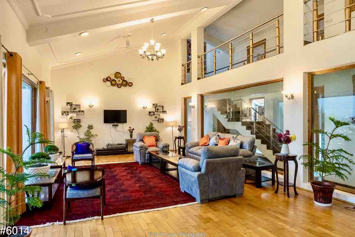 A spacious living area features high ceilings and large windows that allow natural light to enhance the wooden flooring. Comfortable sofas are arranged around a coffee table, complemented by decorative plants. A flat-screen TV is mounted on the wall, and unique wall art adds character to the room.
