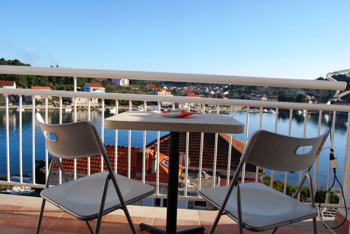 A balcony is presented with a small table and two white chairs, overlooking a serene body of water. The view includes colorful rooftops of nearby buildings, contributing to the peaceful ambiance of the setting.