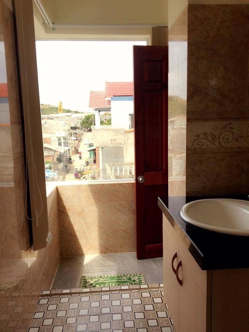Natural light fills a spacious bathroom featuring a sink and countertop made of dark stone. The doorway opens to a balcony area, offering views of the surrounding neighborhood and nearby structures.