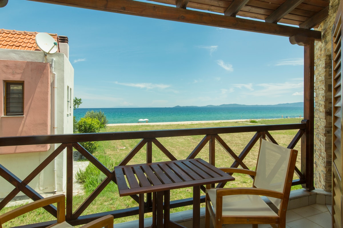 A balcony view showcases the tranquil sea and distant shoreline, framed by a wooden railing. A small table and two chairs are positioned for outdoor relaxation. The sky is bright, reflecting a calm ambiance over the landscape.