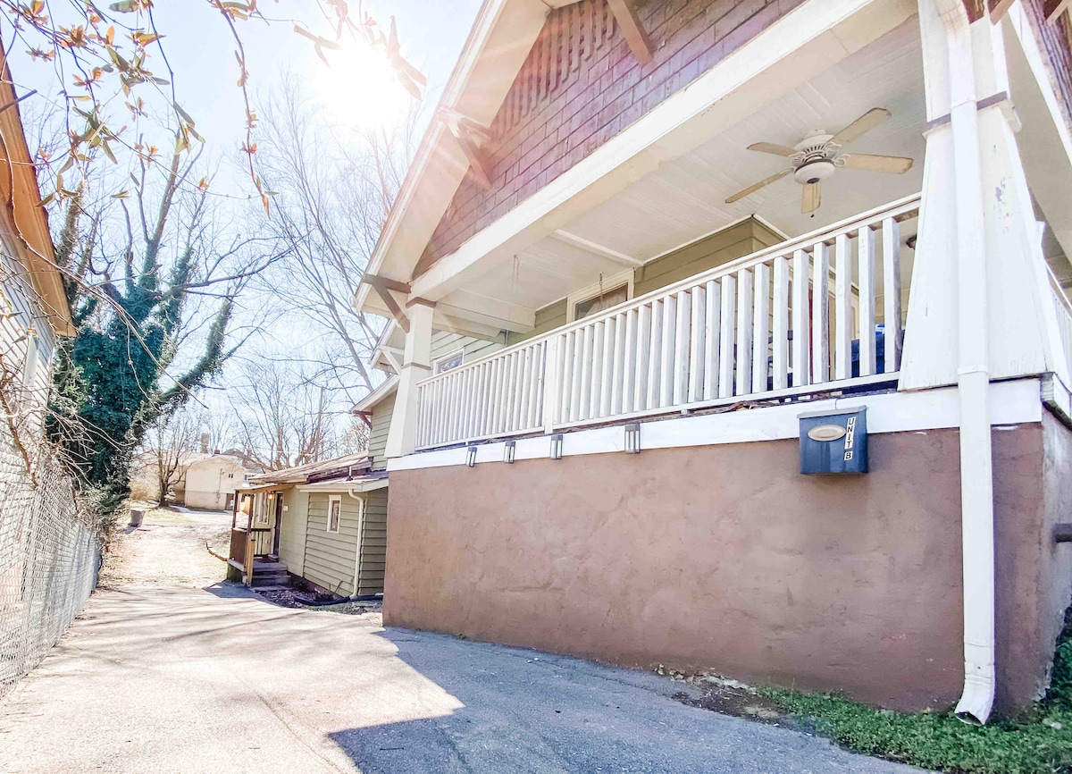 A welcoming porch extends across the front of the home, featuring white railings and a ceiling fan. The entrance is accessible via a paved driveway, which leads to a secondary structure behind the main house. Sunlight reflects off the surface, creating a bright atmosphere.