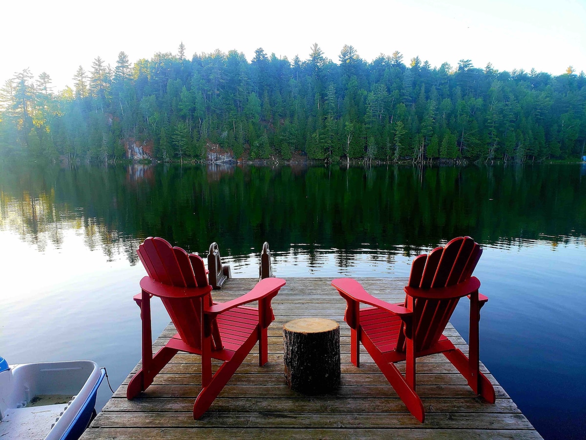 Two red Adirondack chairs are placed on a wooden dock, facing a calm lake surrounded by lush greenery. A small tree stump serves as a side table, and the still water reflects the serene forest backdrop.