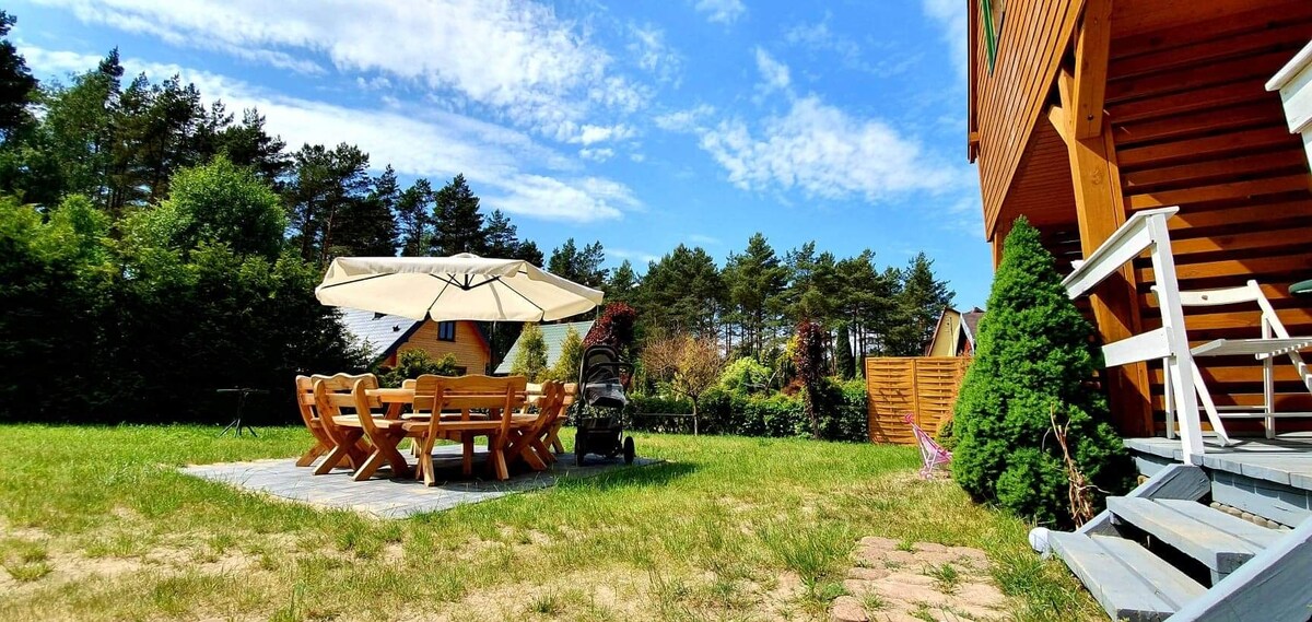 An outdoor seating area is shown with a large wooden table surrounded by multiple chairs, positioned on a stone patio. A shaded area is created by a large umbrella. Lush grass and trees provide a natural backdrop, contributing to a relaxed environment.