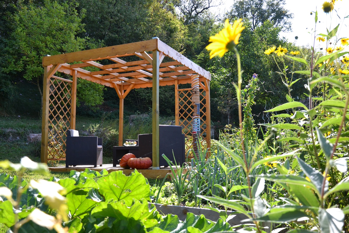 A shaded wooden pergola offers comfortable seating adorned with cushions, surrounded by greenery and blooming flowers in a vibrant garden. The structure features a lattice design, providing a serene space to relax amidst the natural environment, enhancing outdoor enjoyment.