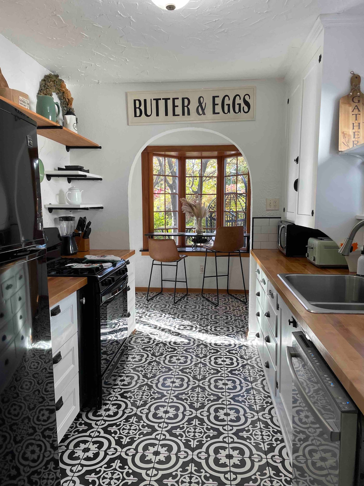 The kitchen features black and white cabinetry complemented by striking patterned flooring. A cozy breakfast nook is visible through an arched window, furnished with two stools. Shelving holds kitchen essentials, and a sign reading 'BUTTER & EGGS' adds a charming touch.