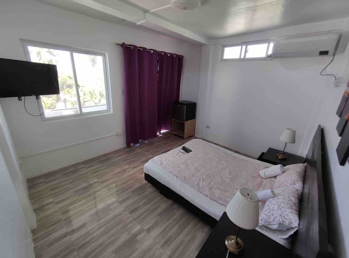 A well-lit room features a spacious bed with floral bedding and soft pillows. A flat-screen TV is mounted on the wall, while a small cabinet sits in the corner. Light filters through the window, with dark curtains offering privacy.