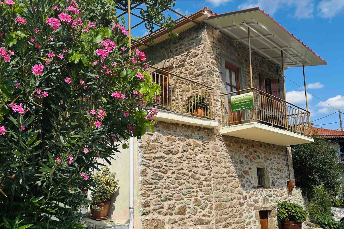 The stone exterior of the holiday home is complemented by vibrant green foliage and blooming pink flowers. A balcony with a railing provides a view, while the roof offers shelter. The surrounding garden adds to the natural setting.
