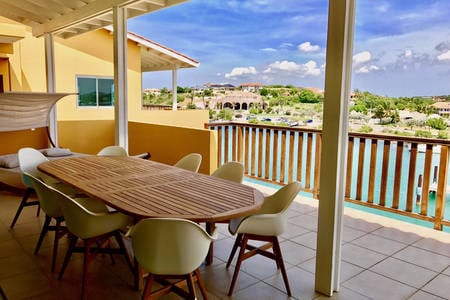 A spacious outdoor dining area is featured, overlooking scenic water views. A large wooden table surrounded by modern chairs offers ample seating. The balcony is framed by light-colored railings, providing an inviting space for gatherings while enjoying the surrounding landscape.