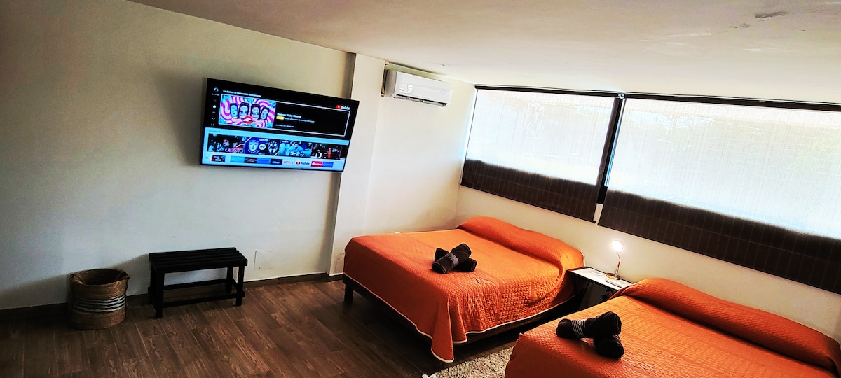 A spacious room features two large beds covered with bright orange bedspreads. A large flat-screen television is mounted on the wall opposite the beds. Natural light enters through expansive windows, complemented by a small nightstand with a lamp, providing a functional and comfortable setting.
