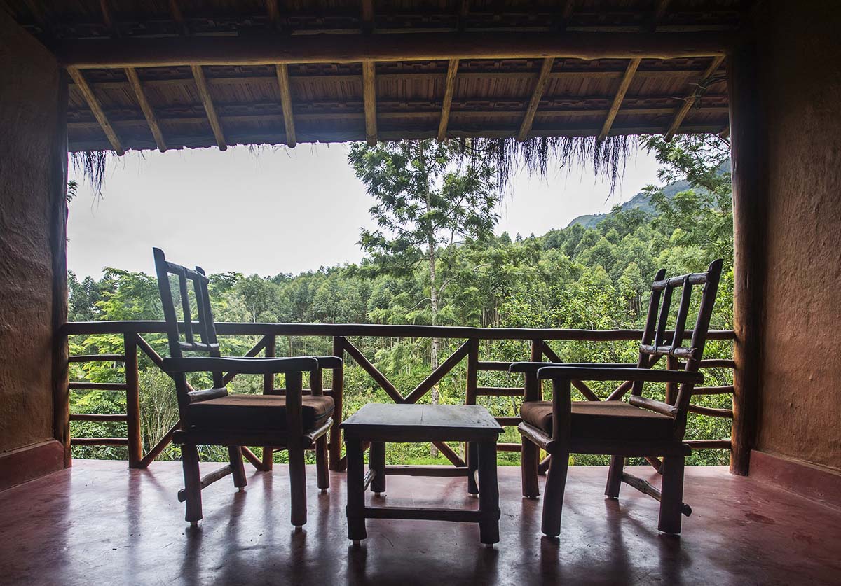 A charming balcony features two wooden chairs and a small table, offering a direct view of the lush greenery and distant hills. The natural setting is framed by a rustic wooden railing and a thatched roof, enhancing the tranquil atmosphere.