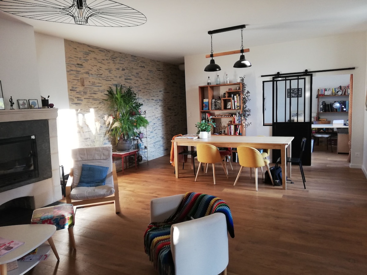 A spacious living area showcases a blend of modern and natural elements. A comfortable armchair is positioned beside a wooden table surrounded by yellow chairs. A stone feature wall complements the light-colored wood flooring, while a large plant adds a touch of greenery.