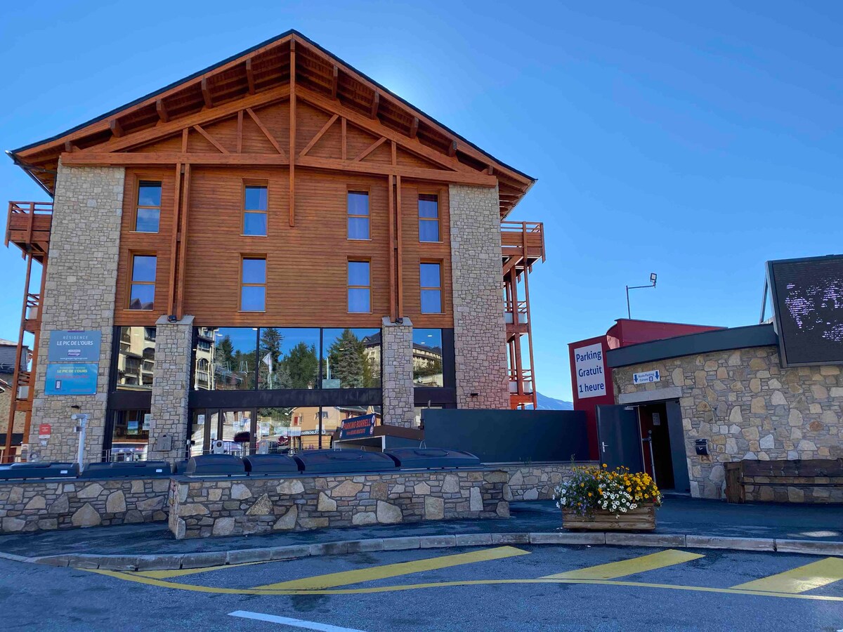 The building features a combination of natural stone and wooden elements, showcasing a rustic yet modern architectural style. Large windows are visible, allowing for ample natural light. The entrance area includes a parking sign and a flower planter with colorful blooms.