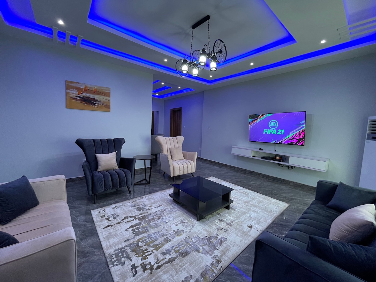 A spacious living area is highlighted by modern furnishings, featuring two armchairs and a comfortable sofa. A large smart TV displays a gaming screen, while recessed lighting in blue accents the ceiling. A patterned rug sits beneath a coffee table, enhancing the inviting environment.