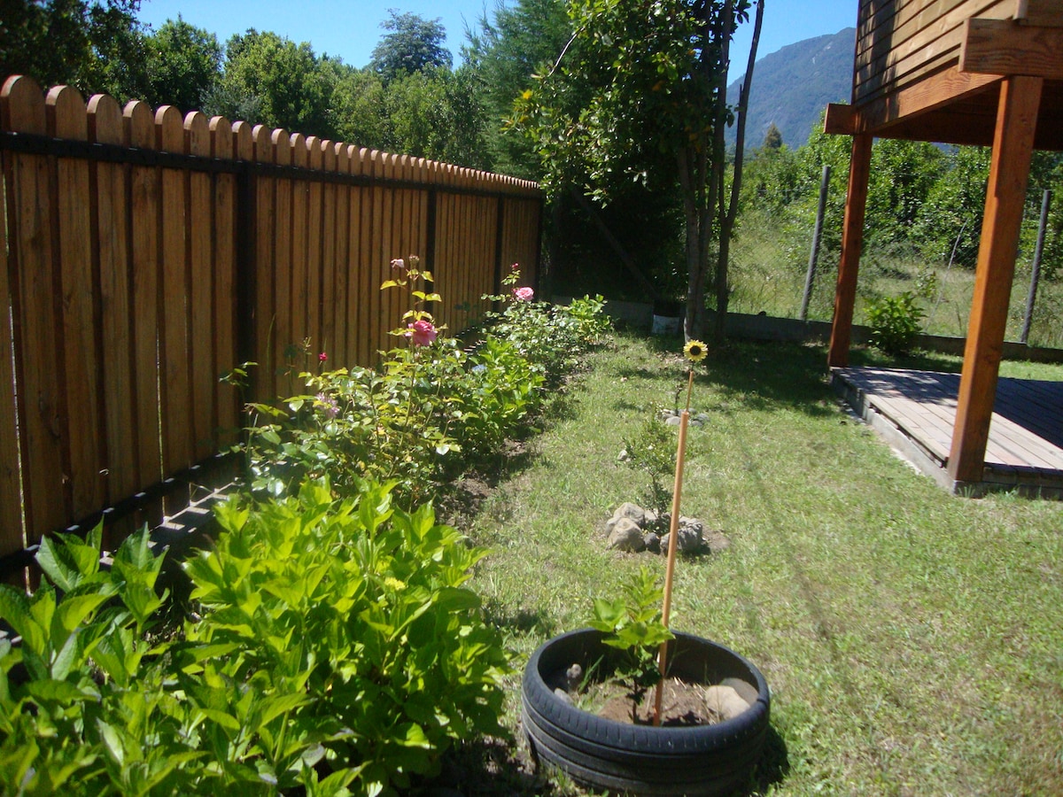 A garden area features a neatly arranged flower bed with various plants, including blooming roses and a newly planted sunflower. A wooden fence lines the property, providing a rustic touch, while the presence of a deck can be seen in the background, set against a mountainous landscape.