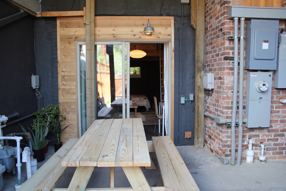 An outdoor covered patio features a sizable wooden picnic table, inviting for al fresco dining. The entrance showcases glass sliding doors that lead into the studio, while natural light filters through, illuminating the rustic wooden walls. A glimpse of the interior can be seen through the doorway.
