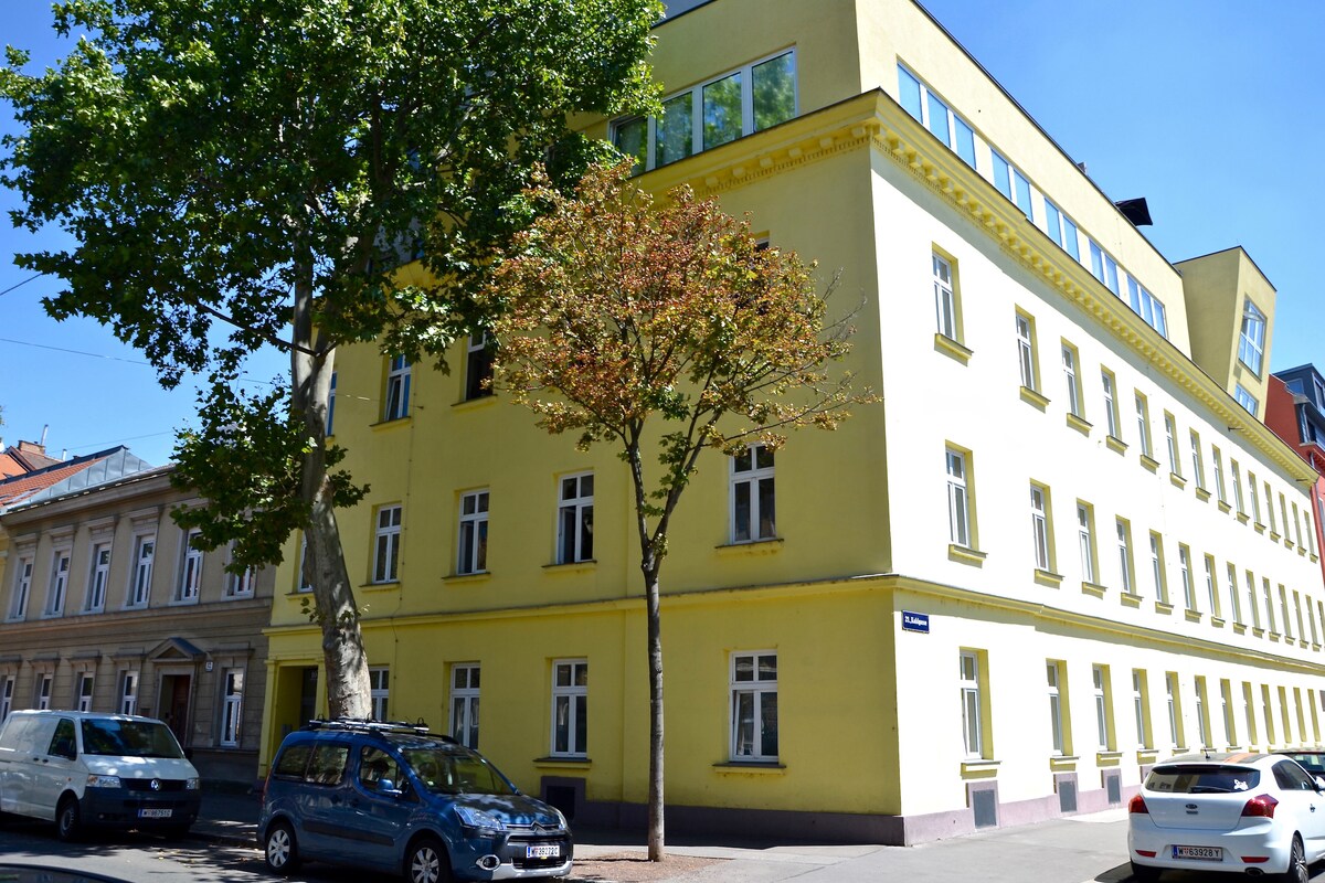 A bright yellow building is presented, featuring multiple windows that allow natural light to enhance the interior. The structure is flanked by trees and vehicles parked along the street, contributing to a welcoming urban environment.