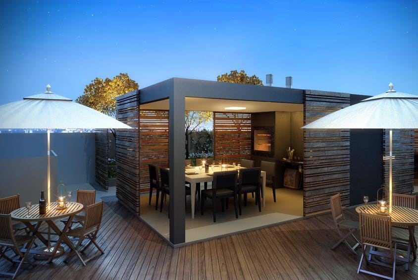 An outdoor dining area features shaded tables surrounded by wooden decking. A modern structure is highlighted by wooden slats and soft lighting overhead. Trees are seen in the background against a twilight sky, creating a relaxed ambiance for social gatherings.