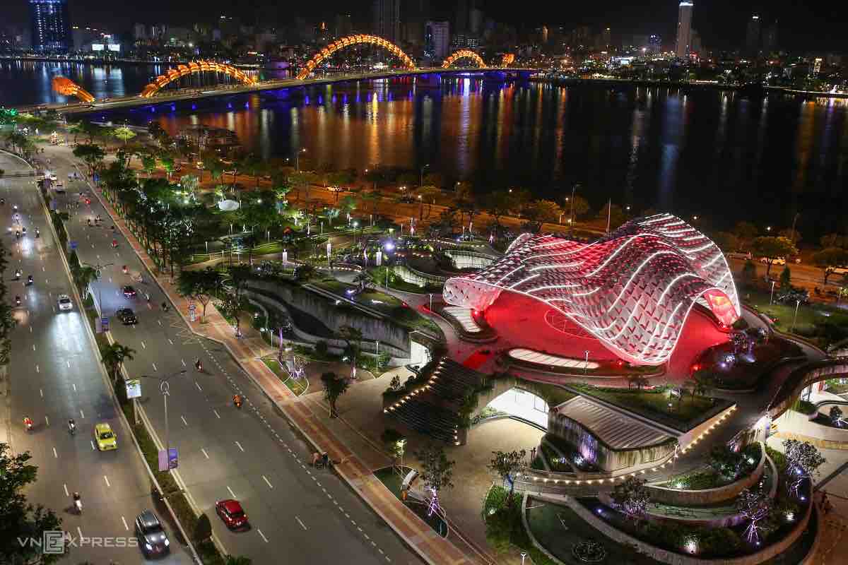 A vibrant riverside scene features the illuminated Rong (Dragon) Bridge spanning the water, with pedestrians strolling along the waterfront. The distinct architectural design of the building, bathed in red light, is prominently displayed amidst lush greenery and softly glowing streetlights, creating a lively evening atmosphere.