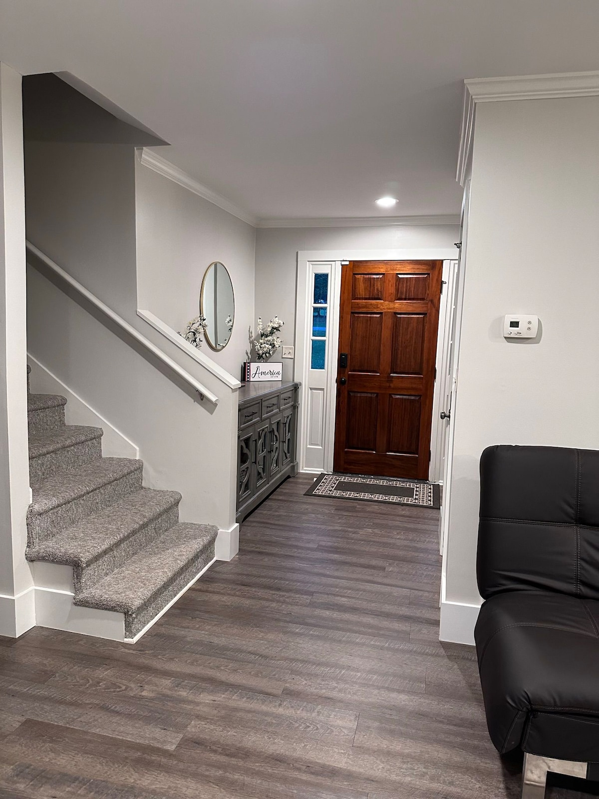 The entrance area features a welcoming foyer with gray walls and a textured staircase leading to the upper level. A console table holds decorative items beside a round mirror, while a wooden front door invites natural light through the window beside it.