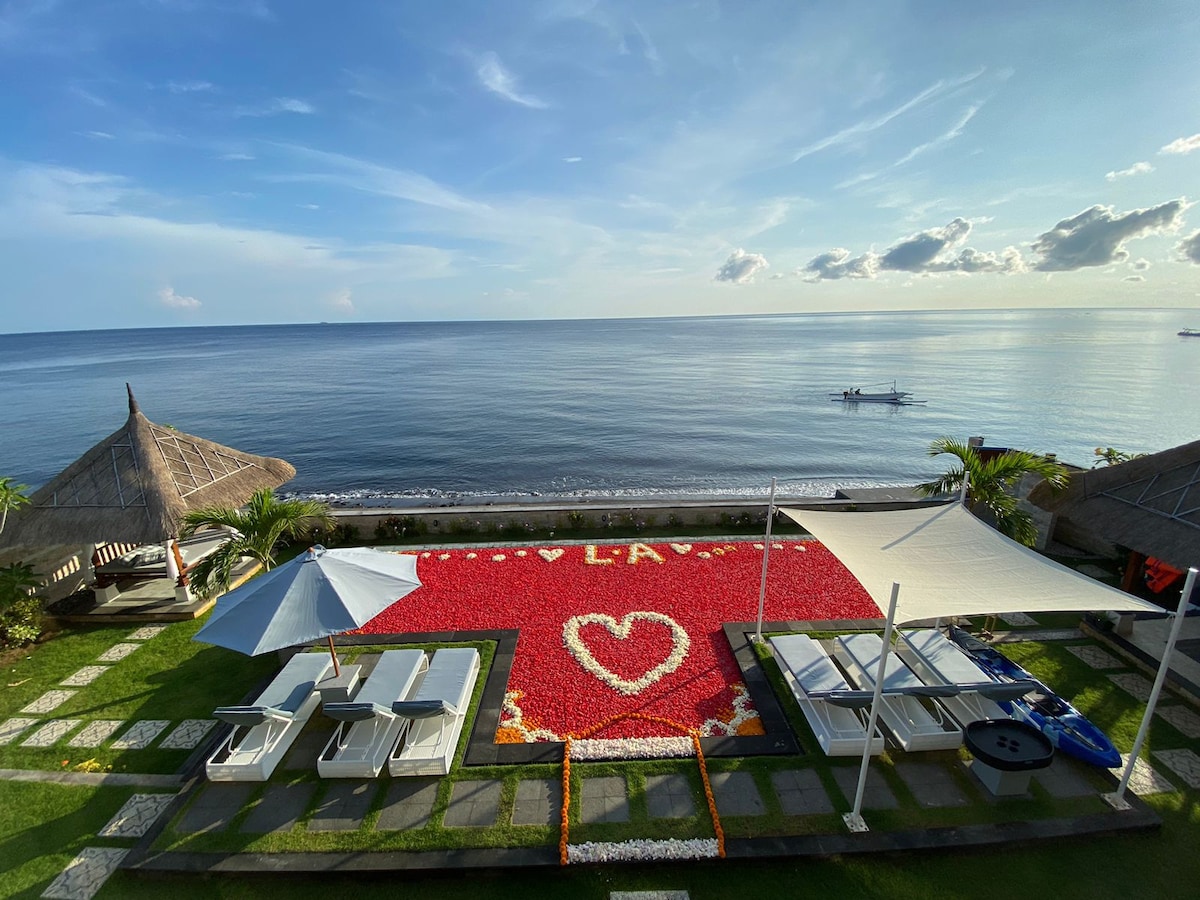 A vibrant beachfront area is decorated with flower petals arranged in a heart shape on the grass. Comfortable lounge chairs are positioned under shade umbrellas, overlooking the calm sea and distant boats. The clear sky enhances the serene atmosphere.