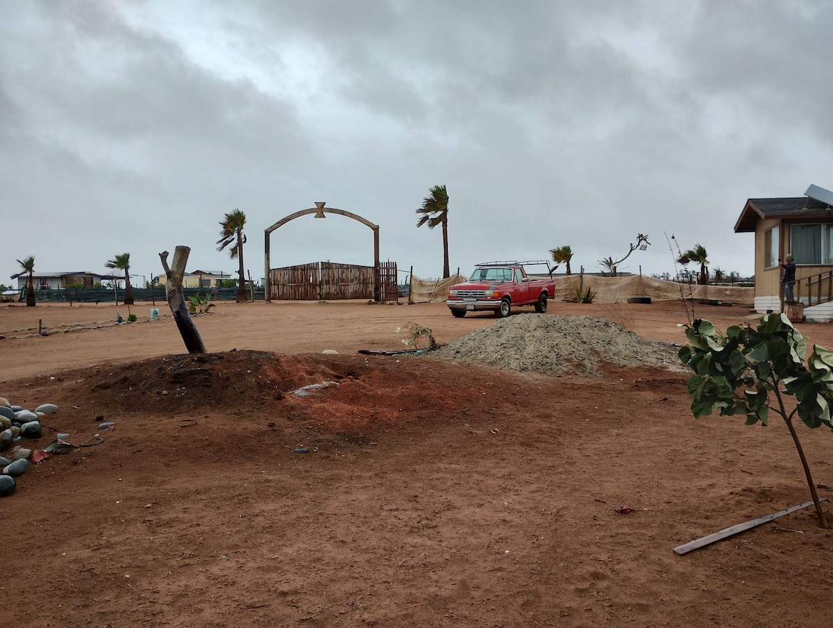 An open area features a gravel path leading toward a wooden gate framed by an arch. A red vehicle is parked nearby, surrounded by sparse landscaping, including scattered rocks and small shrubs. The sky is overcast, adding a calm ambiance to the rural setting.