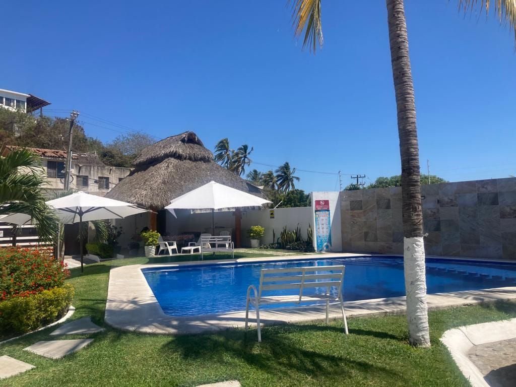 A swimming pool is surrounded by well-maintained grass and palm trees. White lounge chairs and umbrellas provide shaded seating areas. A thatched-roof structure offers additional shelter in the background, contributing to a relaxing outdoor environment.