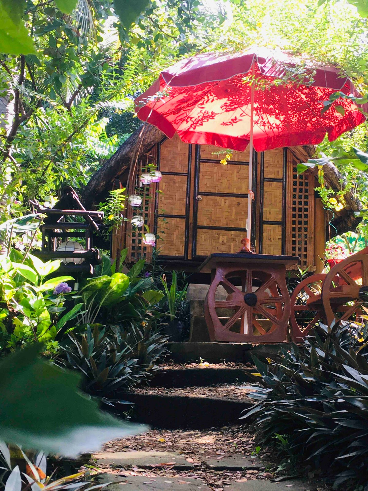 A bamboo kubo is nestled among lush green foliage, accentuated by a vibrant red umbrella. Stone steps lead to the entrance, bordered by decorative plants and wooden wheel accents. Sunlight filters through the surrounding leaves, creating a serene outdoor atmosphere.