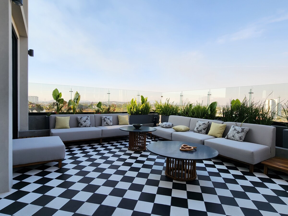 A spacious terrace features modern seating arranged on a checkered black and white floor. Green plants add a touch of nature, while circular tables provide surfaces for drinks and snacks. The backdrop of the city skyline is slightly visible under a clear blue sky.