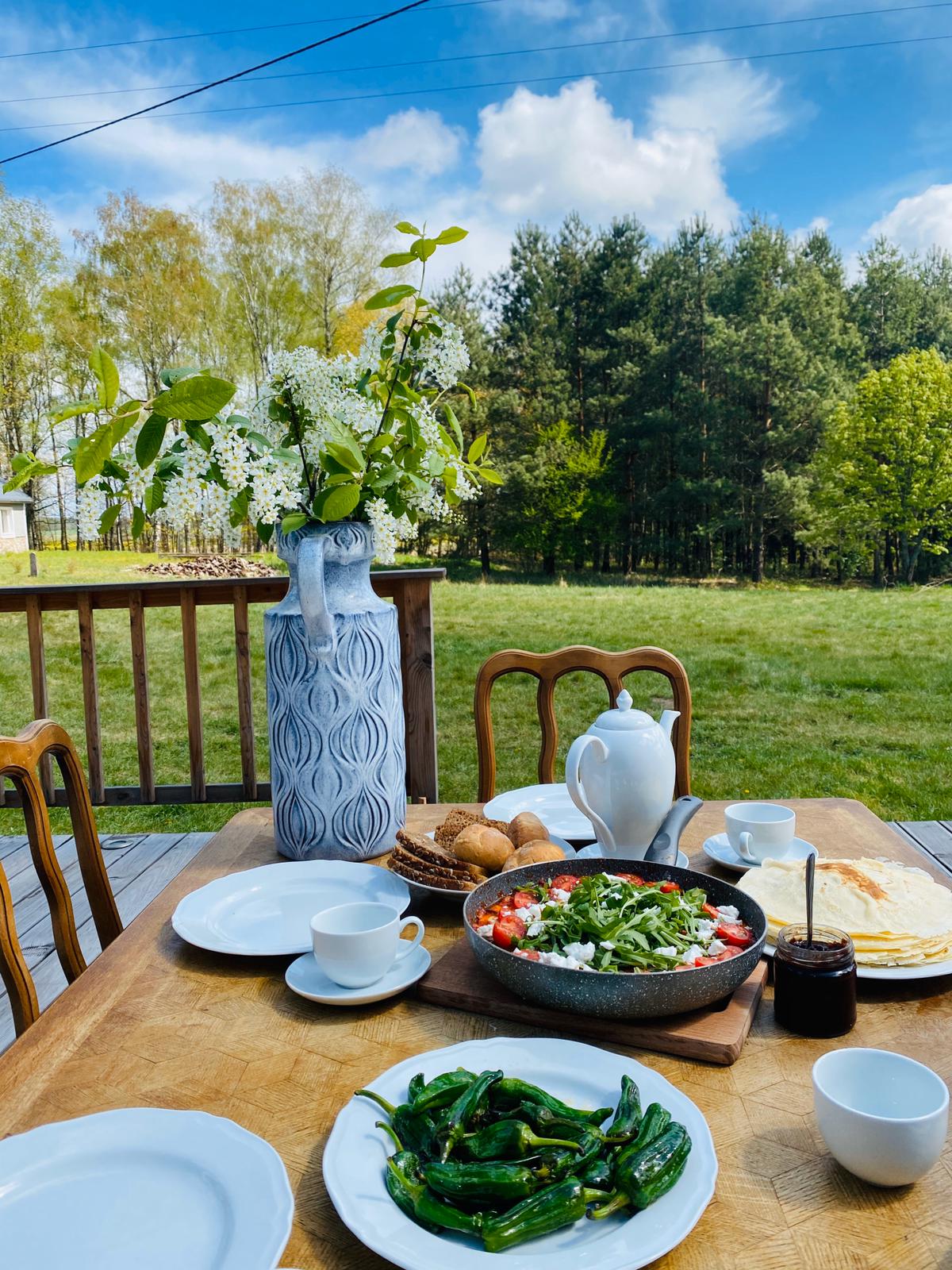 A dining table is set outdoors, featuring plates of fresh salad, grilled vegetables, pastries, and cups beside a teapot. A large floral vase adds a touch of nature, while a green field and trees form a serene backdrop.