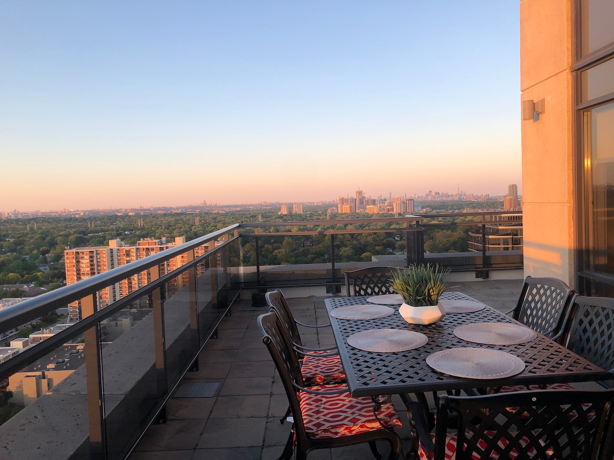 A private terrace is displayed with a dining table surrounded by several chairs. The table is set with six place settings. An unobstructed view of the Toronto skyline is provided, highlighted by the soft hues of sunrise illuminating the distant cityscape.