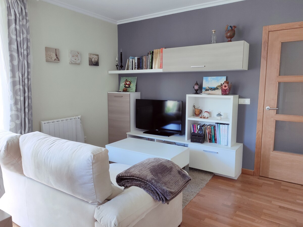 A living area features a neutral-coloured sofa placed in front of a low coffee table. A television is set on a stylish white media unit, surrounded by shelves displaying various decorative items and books. Natural light filters through a nearby window, enhancing the inviting atmosphere.