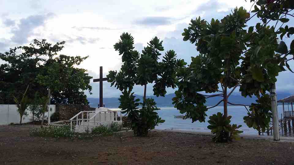 A serene beachfront scene is depicted, featuring lush trees surrounding a cross. The calm ocean is visible in the background under a cloudy sky. A small, white structure is positioned nearby, creating a tranquil atmosphere for reflection and relaxation.