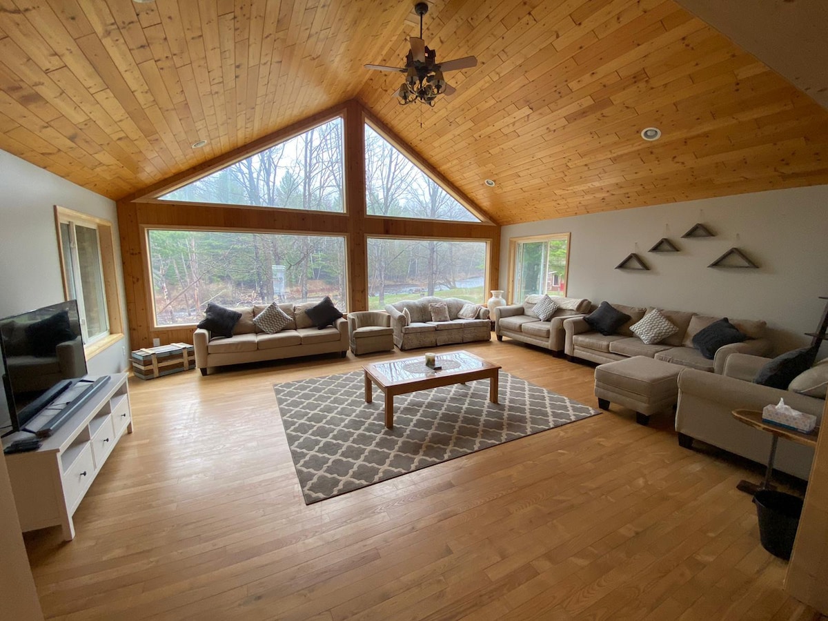 A spacious living room features large triangle windows that frame natural views and invite light. Soft, neutral-colored sofas surround a central coffee table, placed on a patterned area rug. Wooden beams and a ceiling fan complement the warm wood tones of the ceiling.