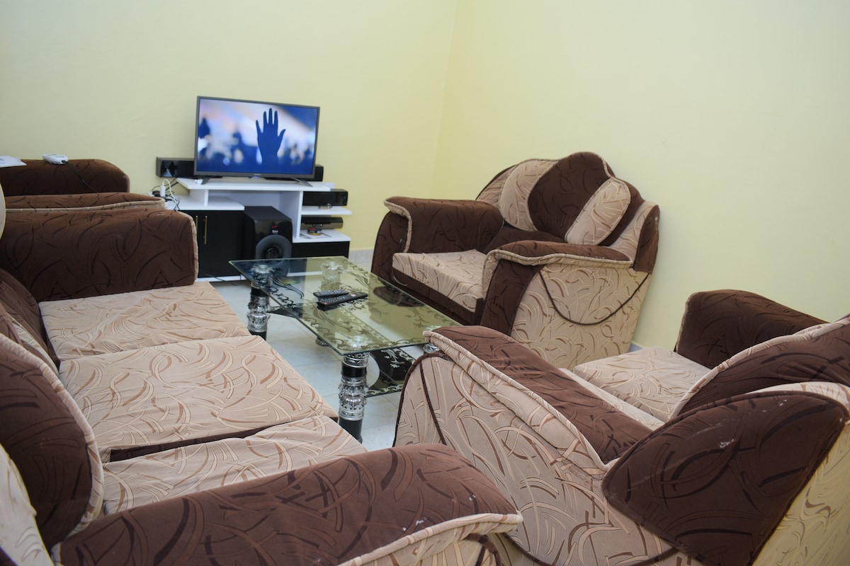 A cozy living room is furnished with a set of comfortable, patterned armchairs and a sofa in earthy tones. A glass coffee table sits in the center, while a television is positioned on a stand against the wall, subtly fitting into the light-colored interior.