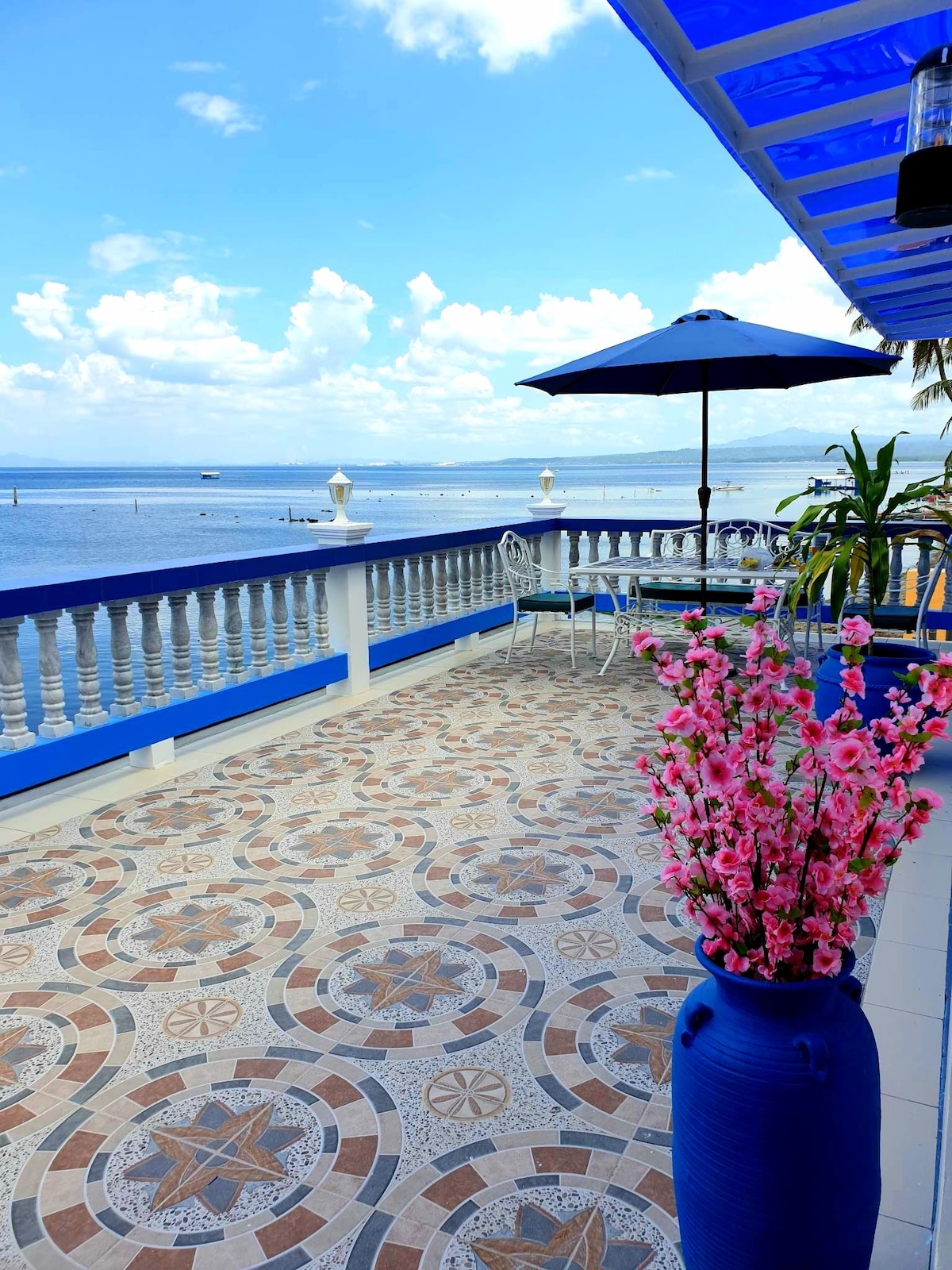 An outdoor terrace features a patterned tiled floor, adorned with a large blue vase filled with pink flowers. A round table and chairs sit under a blue umbrella, overlooking a serene bay and cloudy sky.