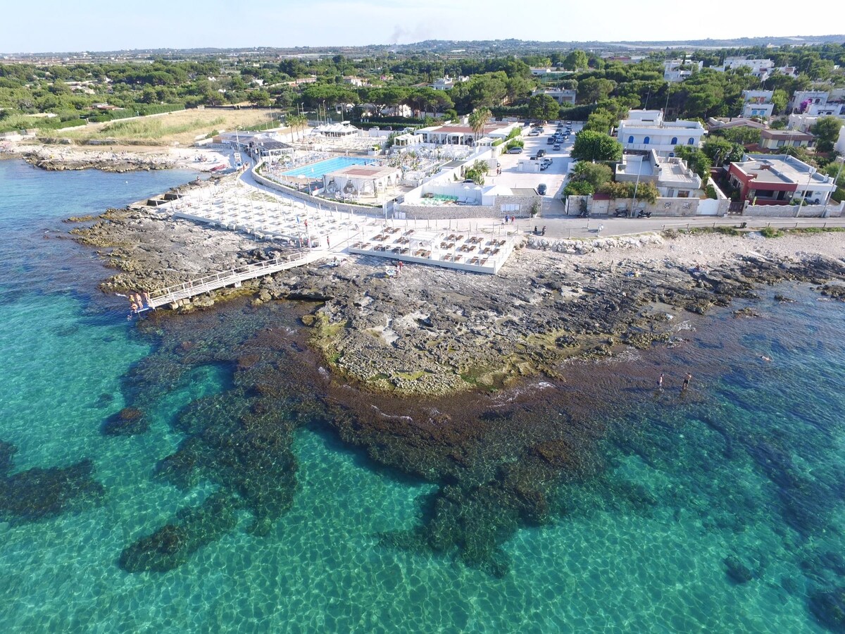 A bird's eye view reveals a serene coastline with clear turquoise waters gently lapping against rocky shores. Sun loungers are arranged for relaxation along the sandy beach, with nearby structures positioned amidst lush greenery, emphasizing a tranquil, inviting atmosphere for visitors.