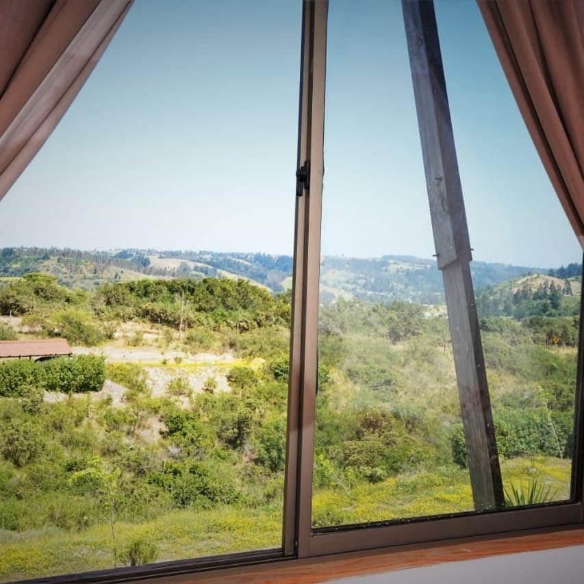 A large window frames a wide view of verdant hills and rolling landscapes under a clear sky. Natural light floods the space, highlighting the surrounding greenery and the peaceful atmosphere, inviting guests to connect with nature.