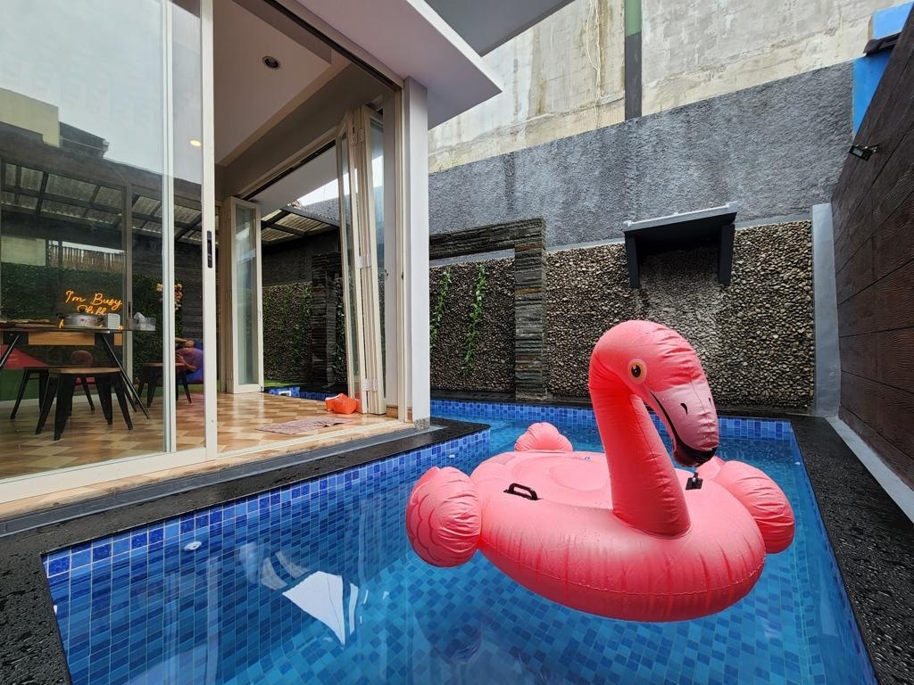 A swimming pool is depicted, featuring a pink inflatable flamingo floating on the water. Surrounding the pool, walls made of stone and greenery create a private atmosphere. Glass doors provide a view of a dining area in the background, enhancing the sense of openness.