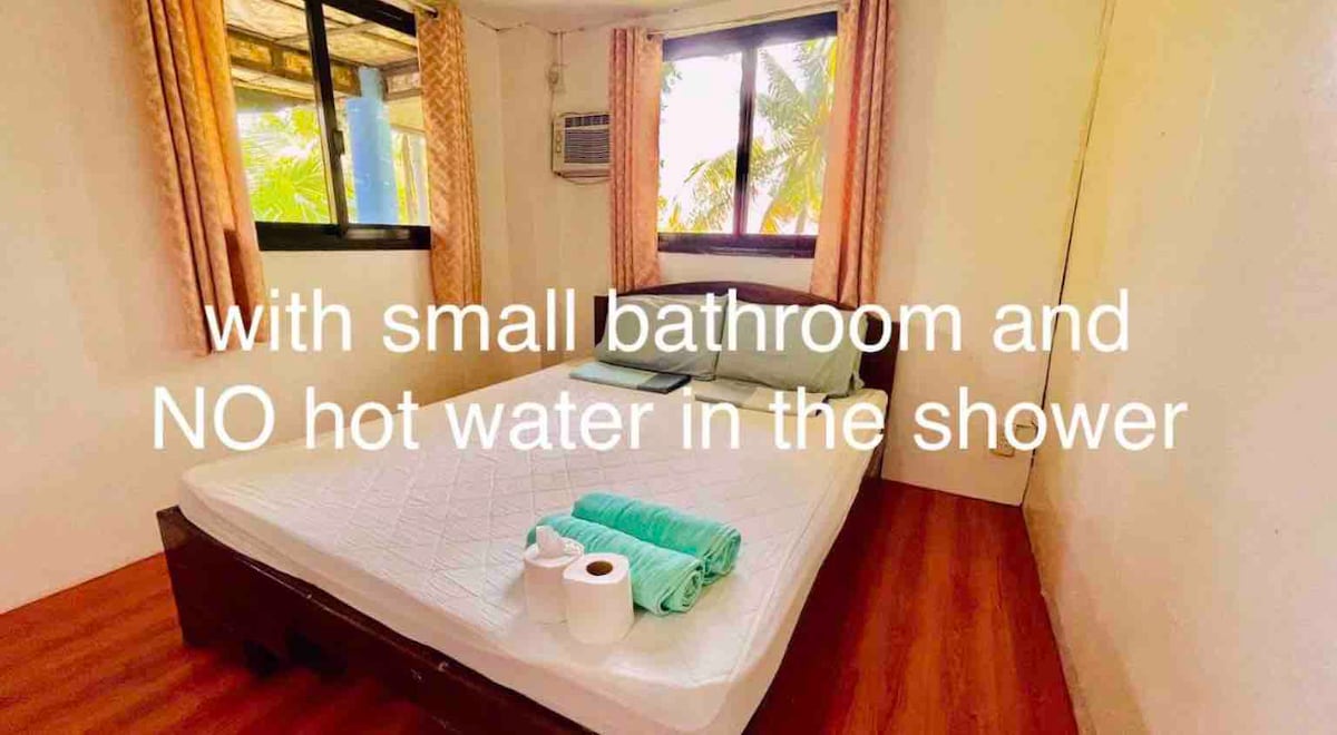 A bright bedroom features a spacious bed with a neatly arranged set of towels. Natural light streams through two windows, framed by soft curtains. A small air conditioning unit is visible, and the flooring is polished wood, contributing to a clean and comfortable environment.