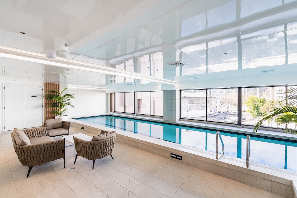 The indoor heated swimming pool is surrounded by large windows that offer natural light and views of the outside greenery. Comfortable seating is provided nearby, with modern furniture arranged to create an inviting space for relaxation.