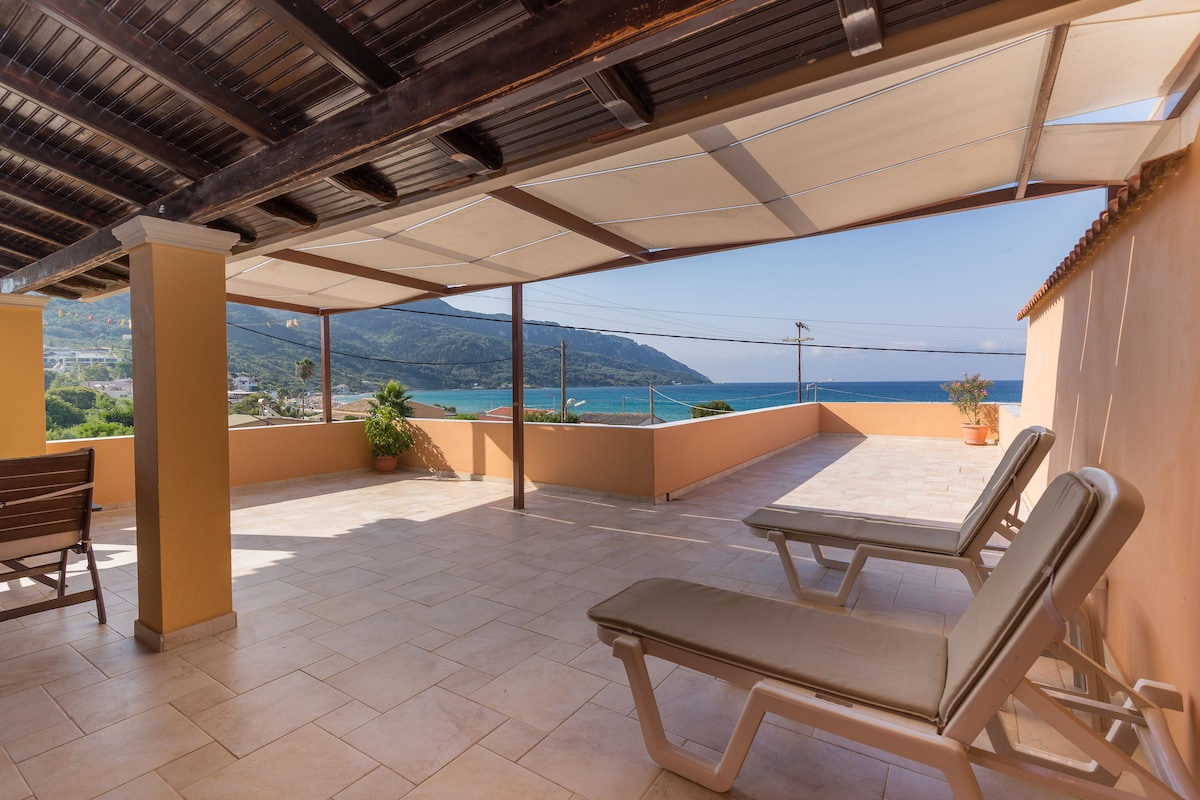 A large terrace offers loungers positioned for relaxation, with a panoramic view of the sea and surrounding landscape. The area is shaded by a canopy overhead, and the tiled floor contributes to a comfortable outdoor setting.