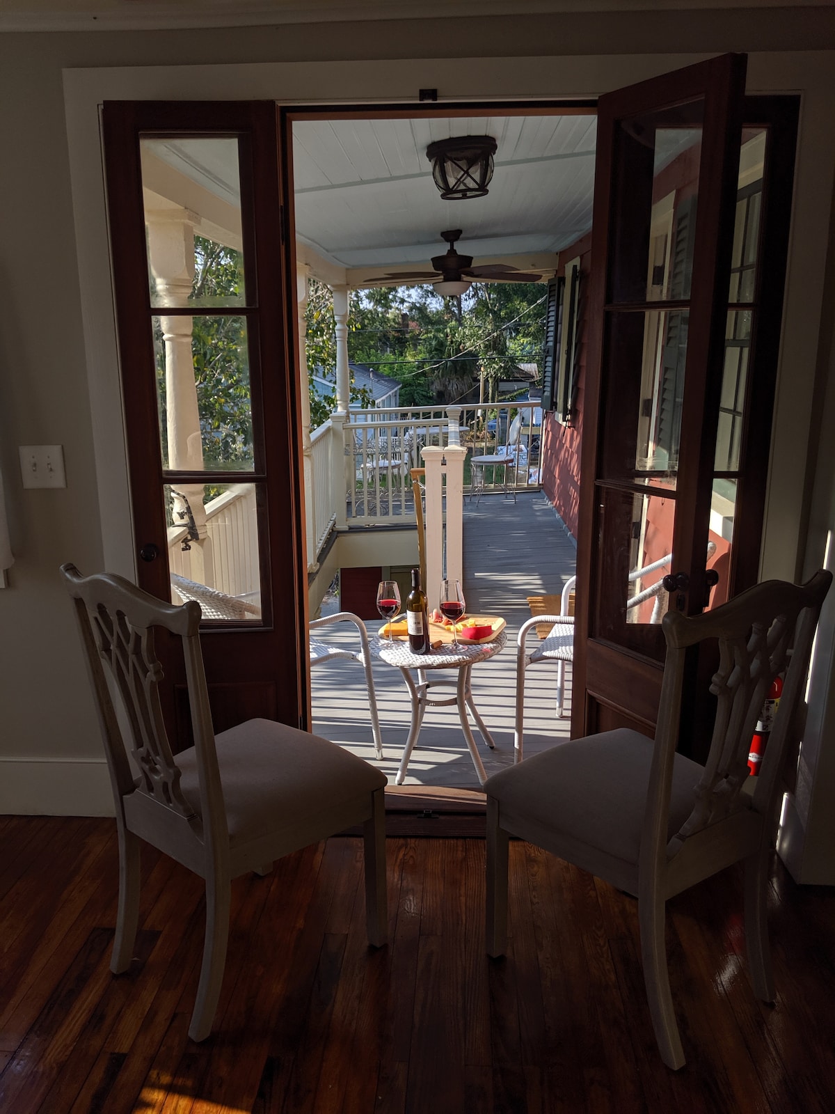A view from inside the apartment shows a pair of elegant chairs facing a small table set with wine glasses and fruit. Double doors lead to a spacious porch surrounded by greenery, inviting enjoyment of the outdoors.