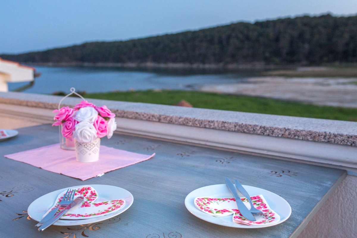 A dining table is set with floral patterned plates and cutlery, complemented by a small decorative vase filled with pink and white artificial flowers. In the background, a serene landscape features a calm body of water bordered by lush green hills.