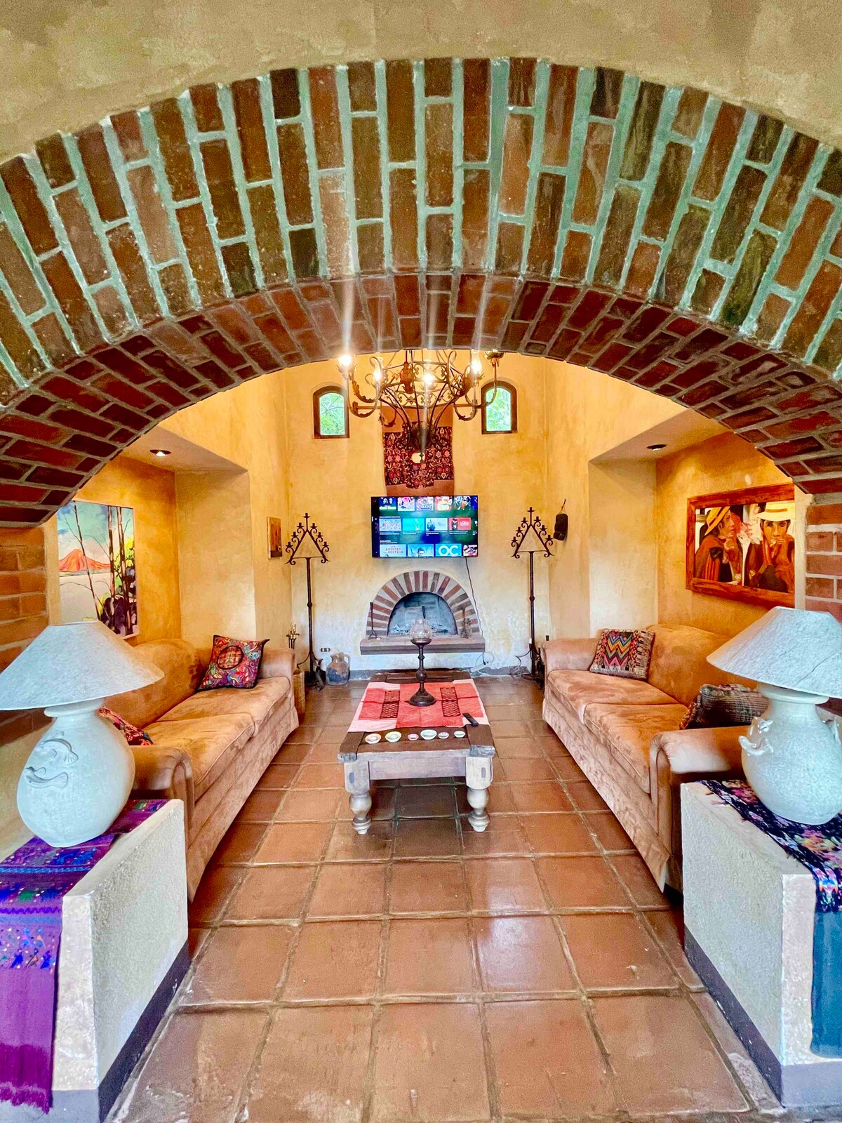 A cozy living area is framed by a rustic brick archway, featuring two light-colored sofas and a central coffee table. A fireplace is nestled between decorative wall sconces, while colorful artwork and a TV adorn the walls. Natural light highlights the terracotta tile flooring.