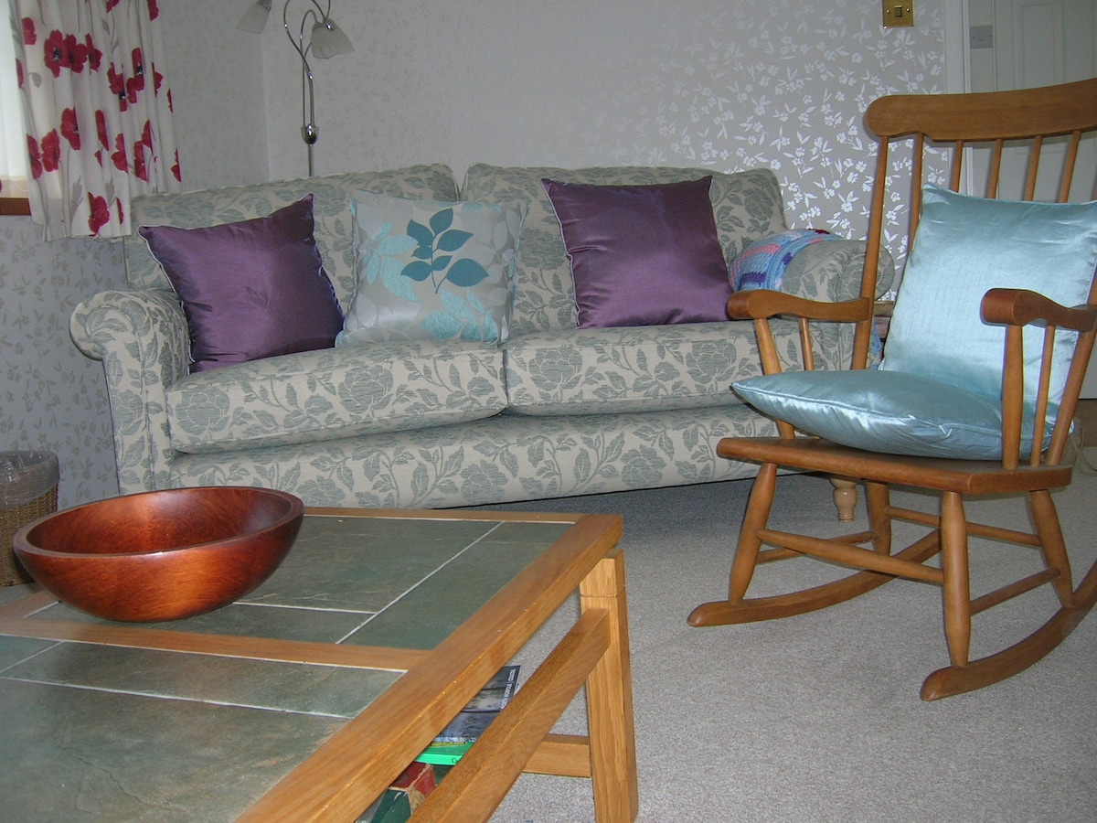 A comfortable seating area includes a floral-patterned couch adorned with purple and teal cushions. A wooden rocking chair is positioned nearby, alongside a square coffee table featuring a decorative wooden bowl, enhancing the inviting atmosphere for relaxation.