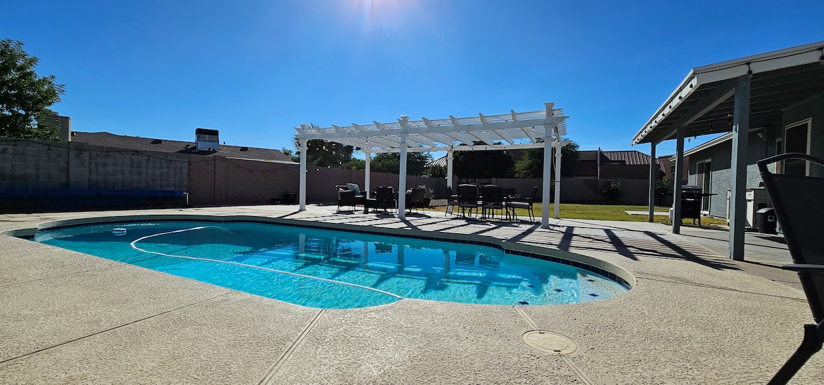 A private heated pool glimmers under the sun, framed by a spacious patio area. A white pergola offers partial shade, while seating arrangements are visible nearby. The surrounding yard features a lush green lawn, providing an inviting outdoor space for relaxation and activities.