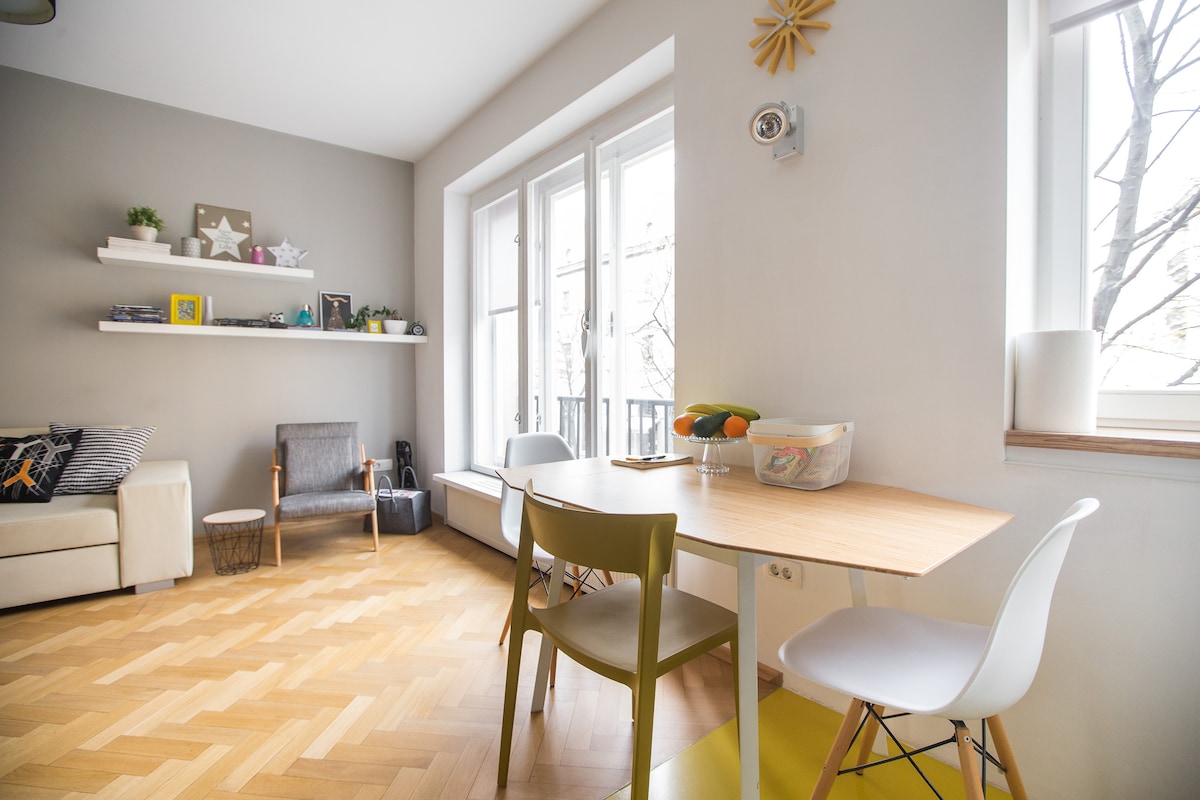A bright and airy living space features large windows with natural light flooding in. A simple dining table is complemented by two chairs. A cozy seating area includes a sofa and an accent chair, with decorative shelves showcasing books and plants.