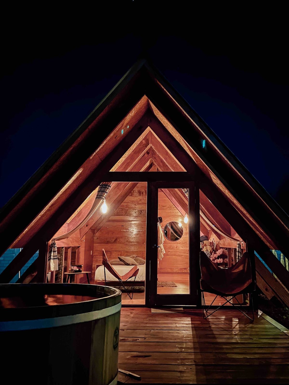 An A-frame cabin glows warmly at night, featuring large glass doors that reveal an inviting interior. Wooden beams create a cozy ambiance. A round hot tub is positioned on the deck, with two chairs set to enjoy the serene environment.