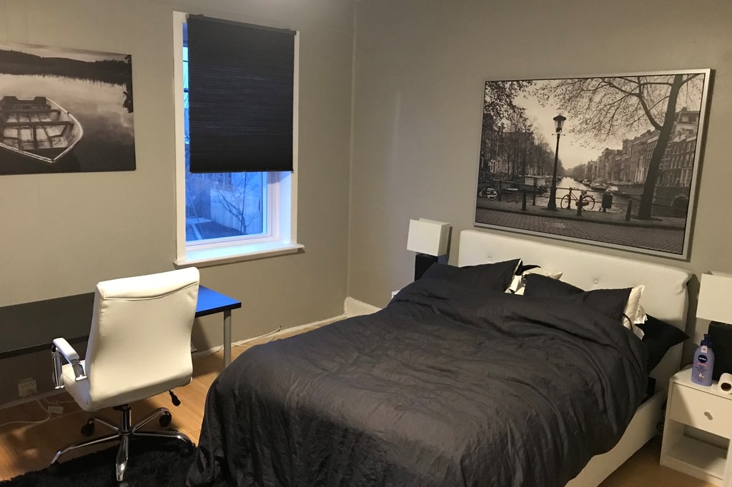 The bedroom features a spacious bed with dark bedding, complemented by a stylish desk and chair. A large window invites natural light, while a black-and-white cityscape photograph adorns the wall. Dark shades provide privacy, enhancing the room's cozy ambiance.