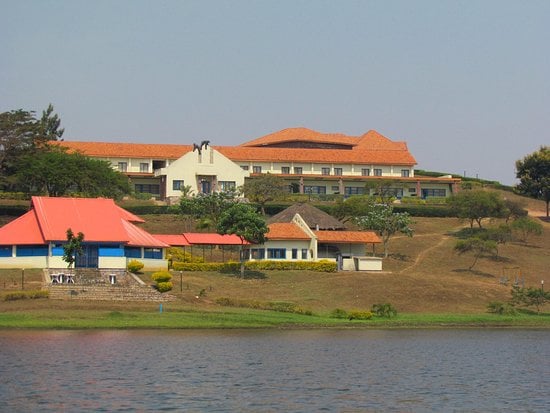 The hotel building is prominently situated on a hill overlooking Lake Muhazi. The structure features a red-tiled roof and multiple levels, surrounded by landscaped gardens and trees. A smaller building with a blue entrance is visible in the foreground, complementing the serene lakeside view.